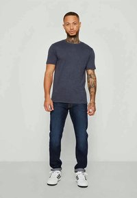 Μπλε μπλουζάκι με στρογγυλή λαιμόκοψη και κοντά μανίκια, συνδυασμένο με σκούρα τζιν και λευκά αθλητικά παπούτσια. Το μοντέλο έχει τατουάζ στους βραχίονες.