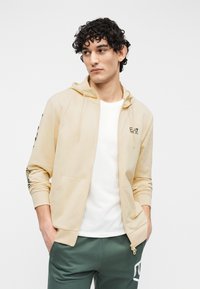 Licht beige hoodie met ritssluiting, voorzakken, verstelbare capuchon en zwarte logodetail. Draagt over een wit T-shirt en gecombineerd met groene broek.