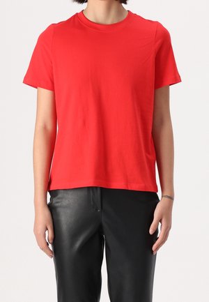 Persoon draagt een effen rood T-shirt met korte mouwen en zwarte leren broek, staand met de armen ontspannen langs het lichaam.