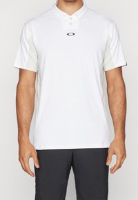 Polo blanc à manches courtes, avec un devant uni et des panneaux latéraux texturés gris clair. Logo noir centré sur la poitrine.