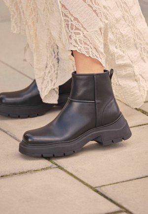 Black leather ankle boots with a chunky sole, featuring a smooth finish and a pull tab on the back. Positioned on a paved surface.