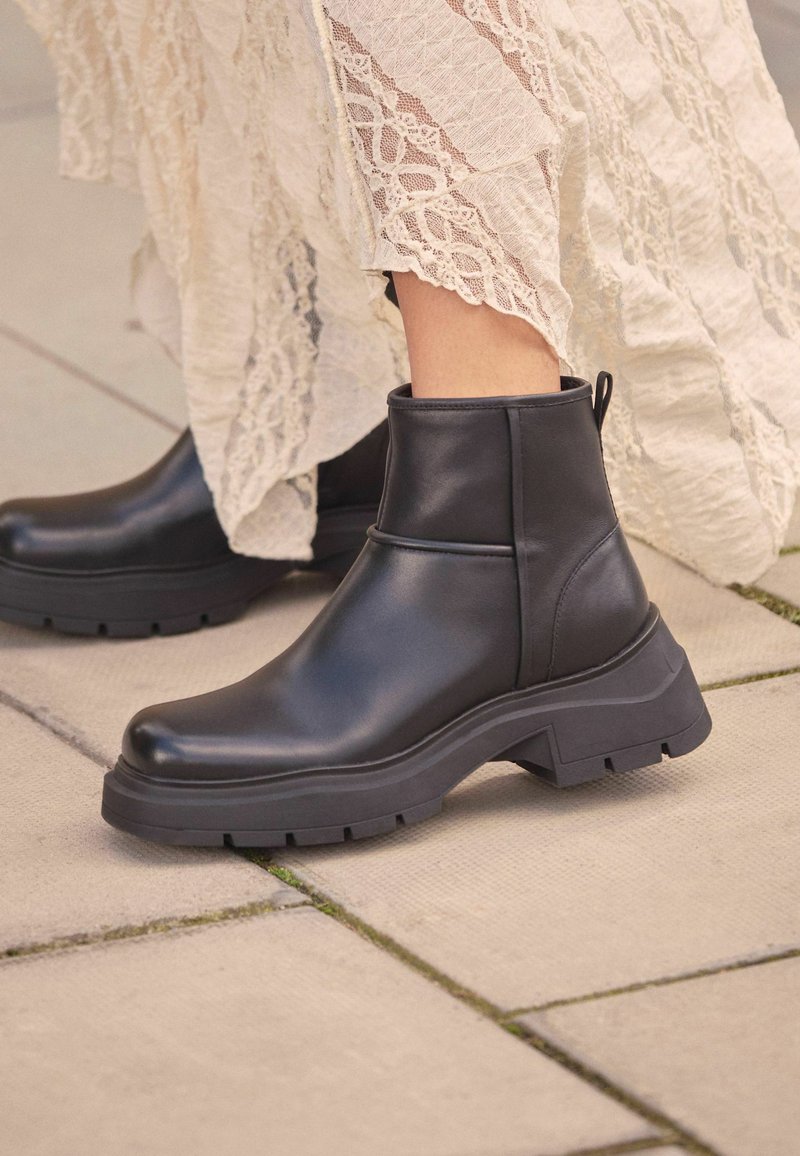 Bottines en cuir noir avec une semelle épaisse, présentant une finition lisse et une poignée à l'arrière. Positionnées sur une surface pavée.