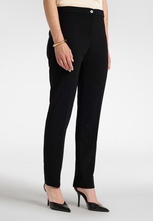 Pantaloni neri aderenti indossati con tacchi alti neri a punta e un braccialetto d'oro sul polso sinistro, mostrati dalla metà della vita fino ai piedi.