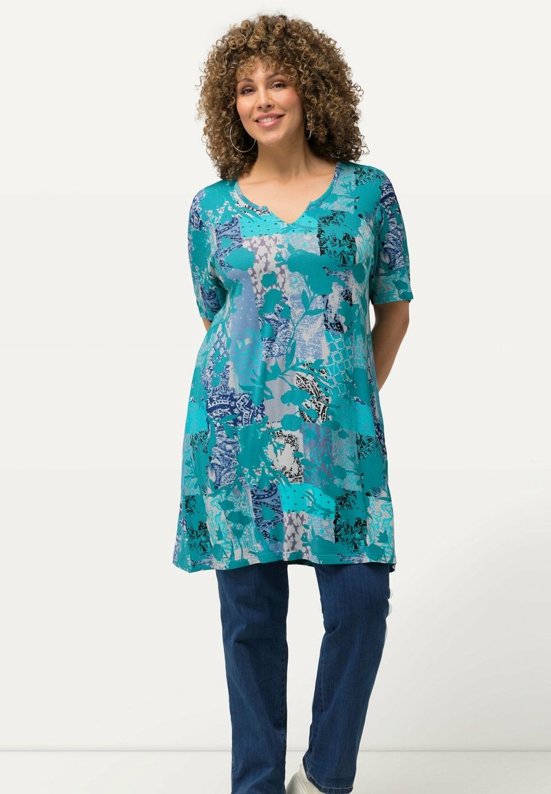 Tunic turchese con maniche corte, con vari motivi astratti in blu, grigio e nero. Abbinata a jeans blu e scarpe bianche.