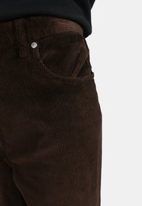 Pantaloni in velluto a coste marroni con una texture a righe, dotati di una tasca frontale e una chiusura a bottone, parzialmente visibili su uno sfondo neutro.