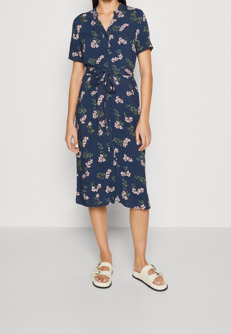 Navy floral dress with short sleeves, button-down front, and a tie belt at the waist. Worn with white sandals featuring decorative hardware.