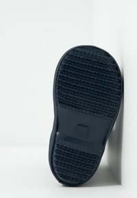 Suela de zapato en goma azul oscuro con una superficie texturizada que presenta surcos y un logo cuadrado. Forma redondeada y bordes limpios.