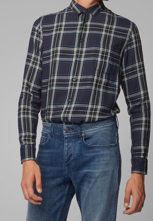 Homme portant une chemise à manches longues à carreaux bleu marine boutonnée, rentrée dans un jean bleu moyen, debout devant un fond gris.