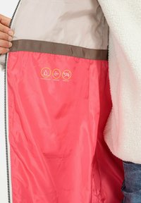 Leichte Jacke mit beiger Außenseite, pinkem Futter und braunen Akzenten; verfügt über Symbole für wasserabweisende, winddichte und daunefreie Materialien.