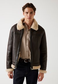 Giacca di pelle marrone con colletto e polsini in shearling beige. Presenta una zip frontale, tasche a toppa e una vestibilità sartoriale. Indossata sopra una camicia leggera.