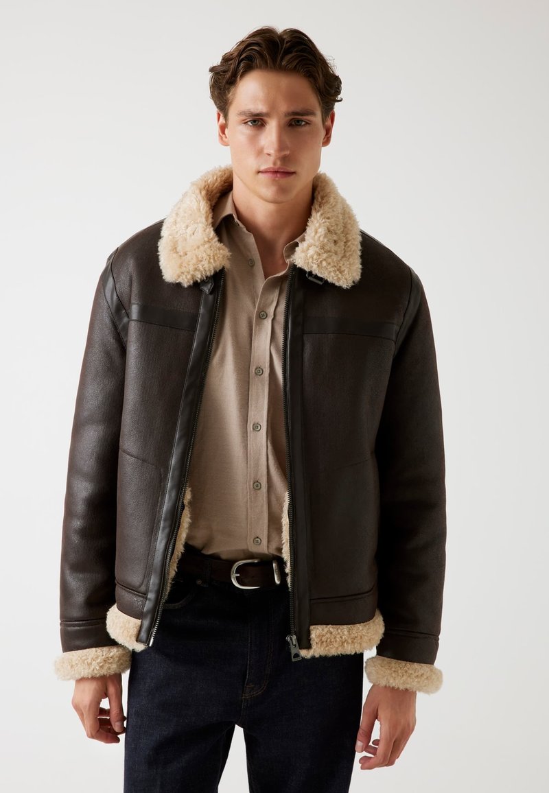 Giacca di pelle marrone con colletto e polsini in shearling beige. Presenta una zip frontale, tasche a toppa e una vestibilità sartoriale. Indossata sopra una camicia leggera.