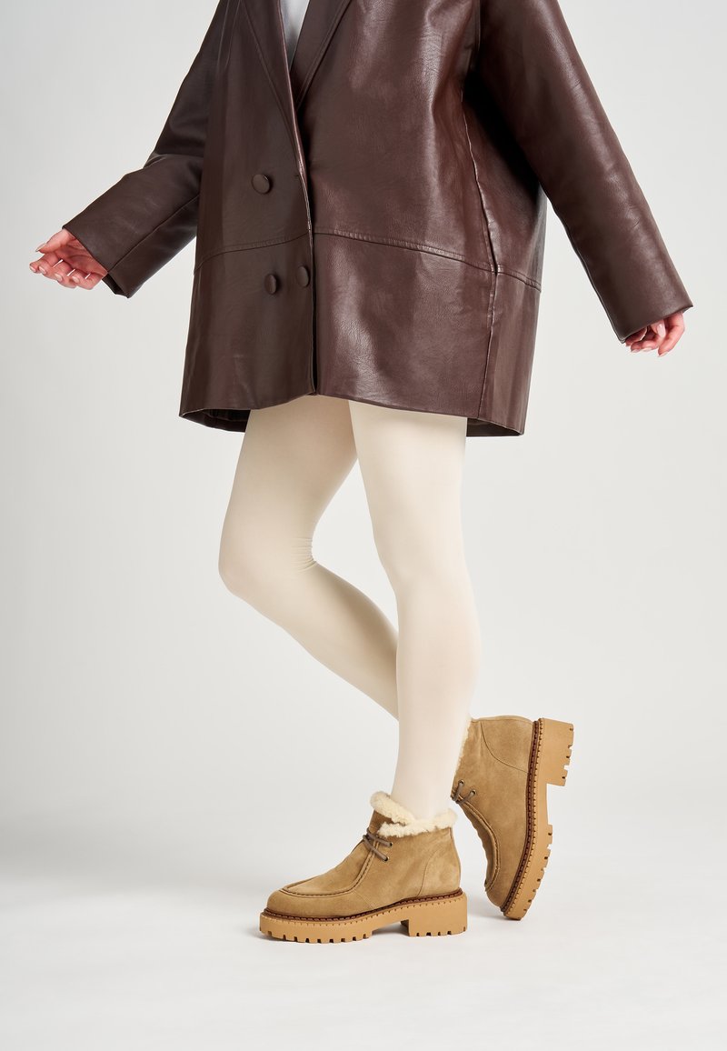 Botas de tobillo de ante marrón claro con forro de piel sintética beige, detalles de cordones y suelas de goma gruesa. Combinadas con medias beige y una chaqueta marrón de gran tamaño.