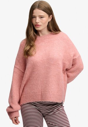 Jonge vrouw met lang bruin haar, die een losse roze gebreide trui en bordeauxrode en witte gestreepte broek draagt, staand voor een witte achtergrond.