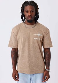 Camiseta de pana marrón con mangas cortas, que presenta un diseño gráfico plateado en el frente. Se lleva con jeans de color azul claro y un collar de cadena.
