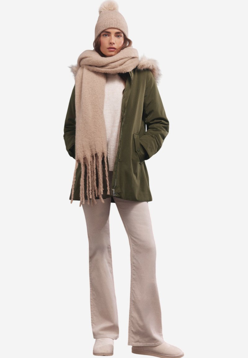 Parka verde con cappuccio bordato di pelliccia, abbinata a una sciarpa beige e un berretto. La modella indossa un maglione color crema e pantaloni chiari, in una posa casual.