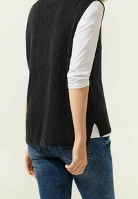 Haut noir sans manches avec une texture côtelée, fentes sur les côtés, porté sur une chemise blanche à manches longues, associé à un jean bleu.