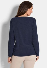 Eine Frau mit langen braunen Haaren, die ein strukturiertes, navyfarbenes Langarmoberteil und eine hellbeige Hose trägt, steht vor einem schlichten weißen Hintergrund.