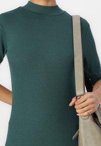 Person wearing a dark green ribbed top with short sleeves, holding a beige leather handbag strap over the right shoulder.