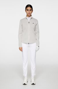 Veste zippée gris clair avec un col haut et des côtés texturés, associée à un pantalon blanc slim et un gant blanc, portée avec des baskets.