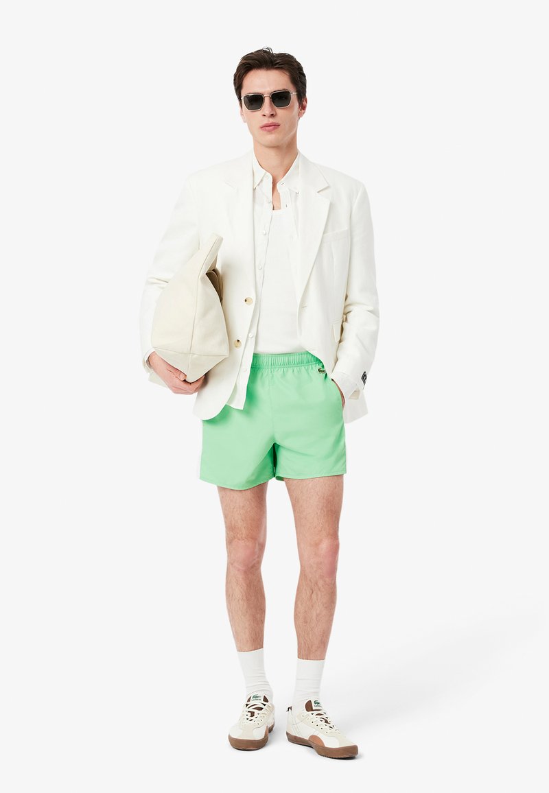 Blazer blanc, chemise blanche, shorts vert menthe et baskets. Les accessoires comprennent des lunettes de soleil et un sac beige. Tissu lisse avec une coupe ajustée.