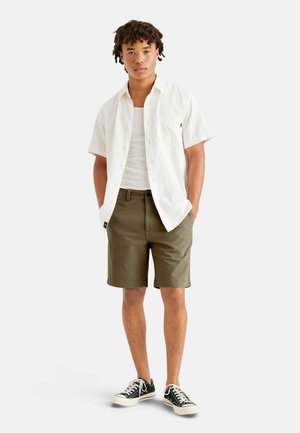 Camisa blanca de manga corta con botones sobre una camiseta de tirantes blanca, pantalones cortos verde oliva y zapatillas negras con suelas blancas. Diseño sencillo y corte relajado.