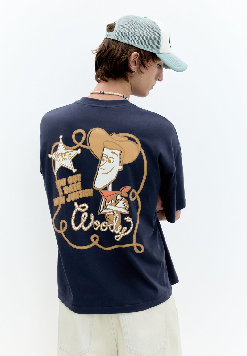 Jeune homme portant un t-shirt bleu marine avec un dessin animé de Woody, un badge de shérif et le texte "You got a date with justice" au dos, casquette claire, pantalon clair.
