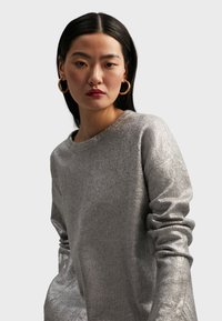 Silverstrukturerad tröja med rund halsringning och långa ärmar, med en subtil glans och rullade ärmslut. Guldörhängen i form av ringar är synliga.