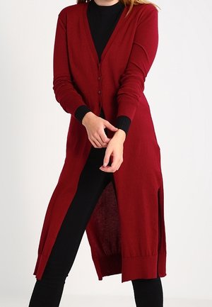 Femme portant un long cardigan rouge boutonné par-dessus un haut noir et un pantalon noir, ajustant la manche du cardigan avec ses mains.