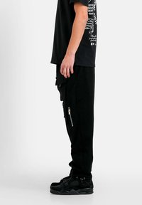Person, der seitlich steht und schwarze Cargo-Hosen mit Reißverschluss, ein schwarzes Grafik-T-Shirt und schwarze Turnschuhe vor einem weißen Hintergrund trägt.