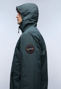 Chaqueta verde oscura con capucha, que presenta un parche circular en la manga. El material parece suave con un acabado mate. Bolsillos laterales con cremallera.
