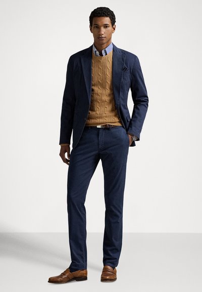 Granatowa marynarka na swetrze w kolorze jasno-brązowym z grubym splotem, do tego granatowe spodnie i brązowe mokasyny, z jasnoniebieską koszulą w paski pod spodem.