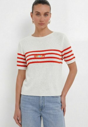 Frau mit dunklen Haaren, die goldene Creolen-Ohrringe, ein weißes Stricktop mit roten Streifen und mehrfarbigem Text sowie hellblaue Jeans trägt.