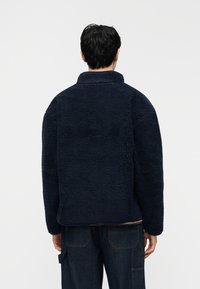 Άτομο που φαίνεται από πίσω φοράει μια υφασμάτινη ζακέτα από φλις σκούρο μπλε χρώμα και σκούρο μπλε τζιν παντελόνια, με απλό ανοιχτόχρωμο φόντο.