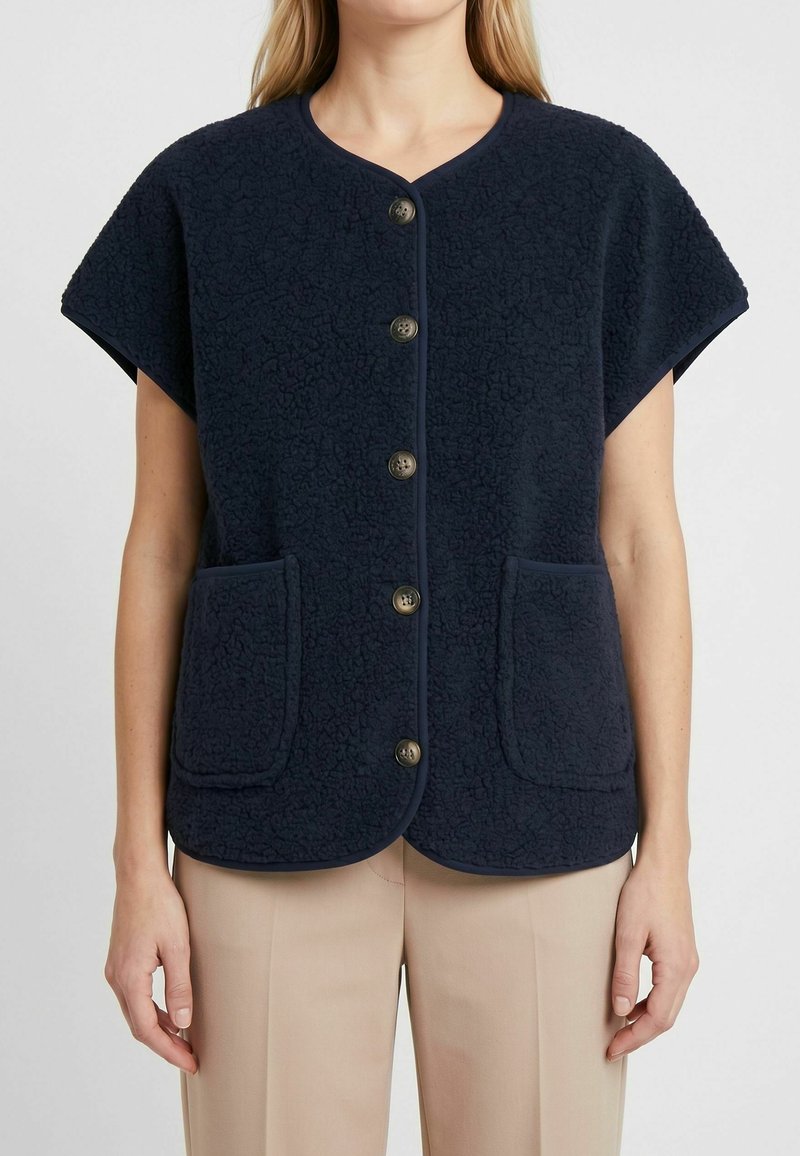 Femme portant un gilet en polaire bleu marine à manches courtes boutonné avec deux poches devant et un pantalon beige, debout devant un fond uni.