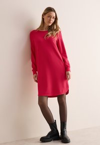 Czerwona sukienka z długim rękawem, okrągłym dekoltem, luźnym krojem i zakrzywionym dołem. Nosi ją w zestawieniu z czarnymi rajstopami oraz stylowymi, grubymi czarnymi botkami za kostkę.