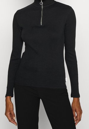 Musta ribbineulottu pitkähiha-paita neljänneksen vetoketjulla ja rengasvetimellä, käytetty mustien housujen kanssa, näkymä keskivartalosta lantioon.