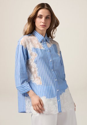 Vrouw draagt een loszittend blauw-wit gestreept overhemd met verticale strepen en beige doorschijnende bloemenkantpanelen, gecombineerd met een witte broek.