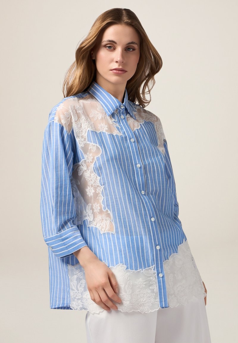 Kobieta ubrana w luźną koszulę z pionowymi niebiesko-białymi paskami i beżowymi przezroczystymi panelami z kwiatowej koronki, w połączeniu z białymi spodniami.