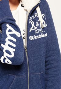 Felpa con zip blu realizzata in tessuto morbido, con un cappuccio foderato in pile bianco, scritte stampate sulle maniche e tasche frontali.