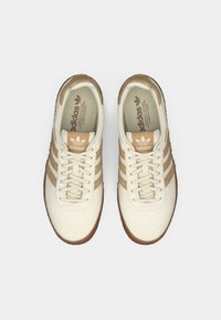 Μπεζ δερμάτινα αθλητικά παπούτσια με τρεις καφέ ρίγες, διάτρητο μπροστινό μέρος, επίπεδα κορδόνια και λαστιχένια σόλα. Λογότυπο "adidas" στη γλώσσα σε ταμπά χρώμα.