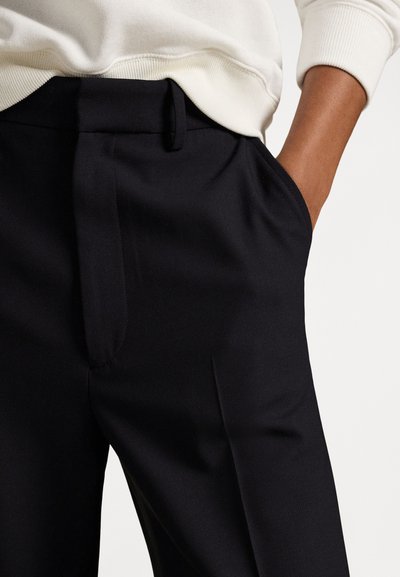 Mão direita no bolso de calças pretas de alfaiate usadas com camisola branca de malha.