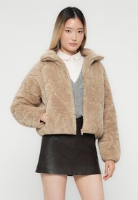Ung kvinde iført en beige, fuzzy zip-up jakke med hjerte-mønstre, grå sweatervest, hvid skjorte med krave og sort læder mini nederdel.