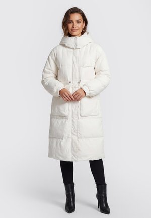 Vrouw draagt een lange witte gewatteerde jas met capuchon, zwarte leggings en zwarte enkellaarsjes met hak en staat met de handen gevouwen voor zich.
