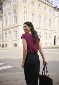 Jeune femme souriante, marchant au coin d'une rue de la ville, portant une chemise bordeaux et une jupe noire, tenant un sac fourre-tout noir, avec un bâtiment historique en arrière-plan.