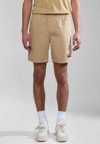 Beige shorts med elastisk talje, sidelommer, flad søm og et patchlogo på det venstre ben, parret med hvide sneakers og sokker.