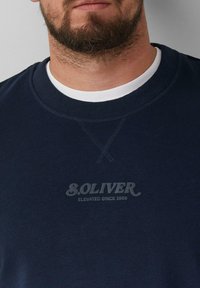 Felpa azul navy realizzata in tessuto morbido, con un colletto rotondo a coste e una stampa logo testurizzata che recita "S.OLIVER ELEVATED SINCE 1969."