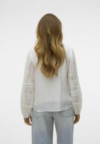 Blusa bianca a maniche lunghe con corpo testurizzato, dettagli in pizzo sulle maniche e polsini con bottoni, abbinata a jeans azzurri chiari.