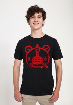 Ung mand med krøllet hår smiler og bærer en sort T-shirt med en stor rød bamseansigtsgrafik med spænder og stropper.