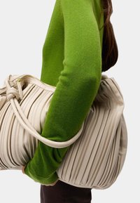 Cremeväskan med veckad design och texturerad yta, hålls av en person i grön tröja. Den har ett knutet handtag och dragkedjestängning.