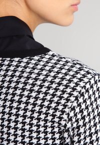 Svart och vit houndstooth-mönstrad tröja med svart krage. Stickad struktur med passande form, som framhäver designens detaljer.